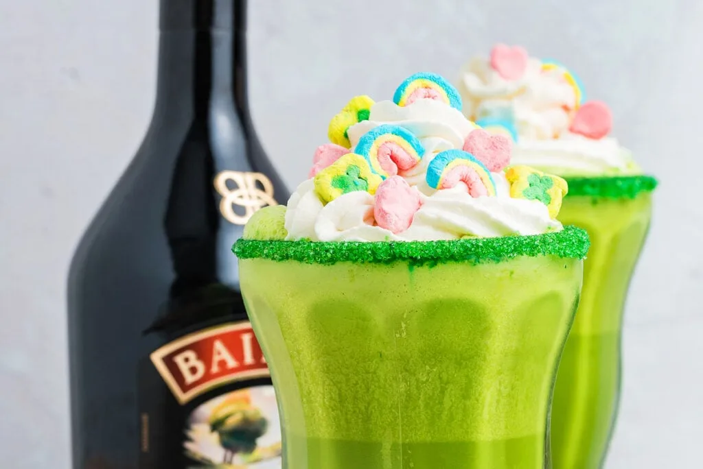 A vibrant green cocktail topped with whipped cream, colorful marshmallow rainbows, and a sugar-rimmed glass, with a bottle of Baileys in the background, creating a festive and indulgent green drink.