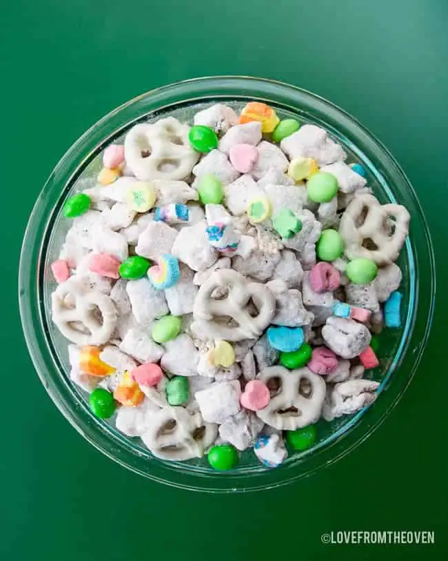 A festive bowl of St. Patrick's Day snack mix featuring white chocolate-covered pretzels, green candies, colorful marshmallows, and powdered sugar-coated treats for a sweet Irish party food!