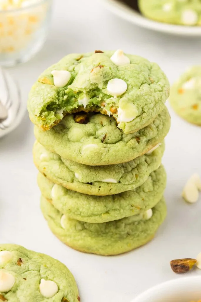 A stack of soft green pistachio cookies with white chocolate chips, with the top cookie partially eaten, perfect for St Patrick's Day desserts or Irish-inspired treats.