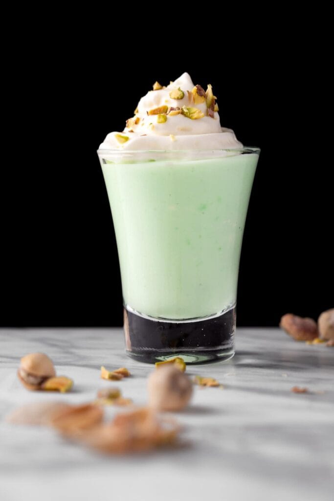 A creamy green dessert drink served in a small glass, topped with whipped cream and chopped pistachios, placed on a marble surface with scattered pistachios in the foreground and a black background for contrast.