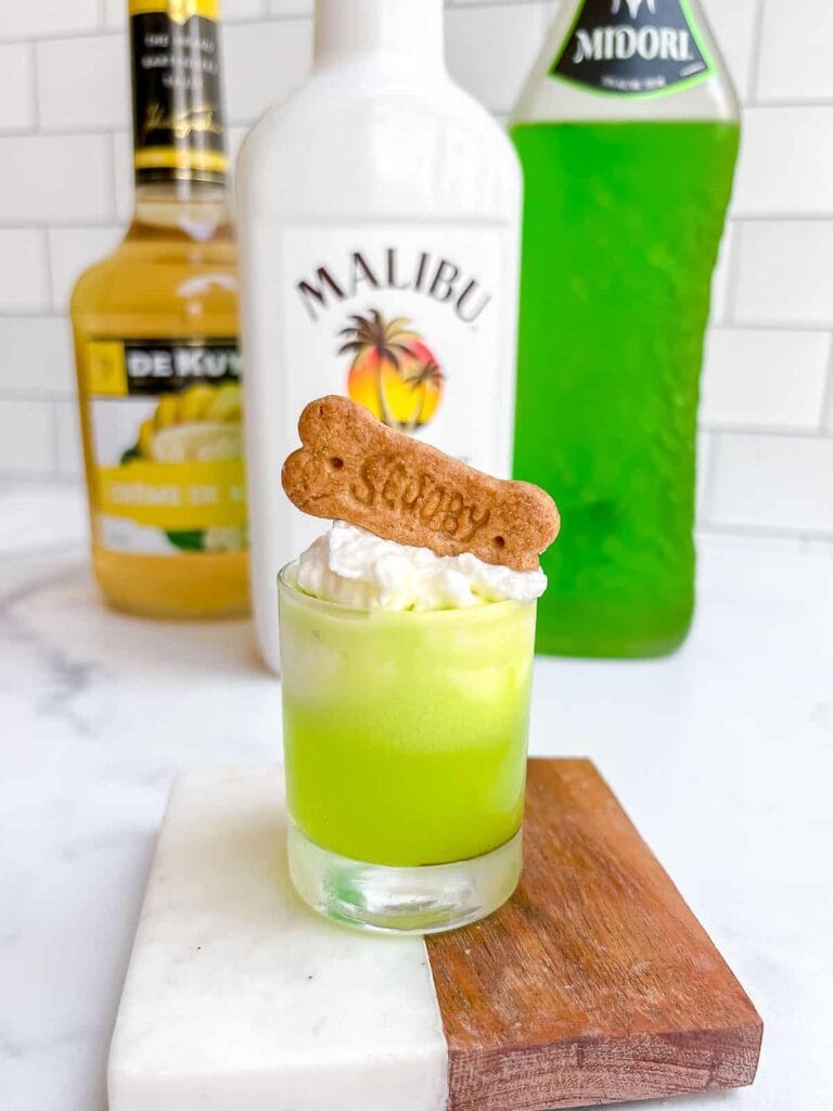 A fun green shot topped with whipped cream and a Scooby-Doo-themed cookie garnish, set on a marble and wood serving block, with bottles of Malibu, Midori, and pineapple liqueur in the background.