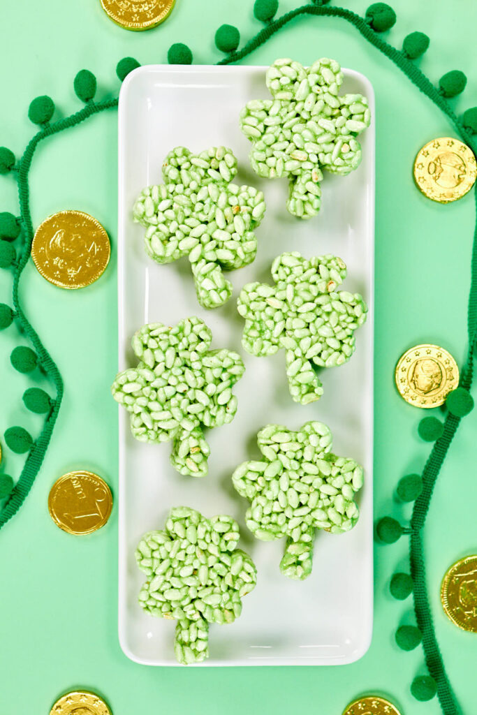 Shamrock-shaped green rice crispy treats arranged on a white tray, surrounded by gold chocolate coins and festive green decorations, perfect for St. Patrick's Day celebrations.