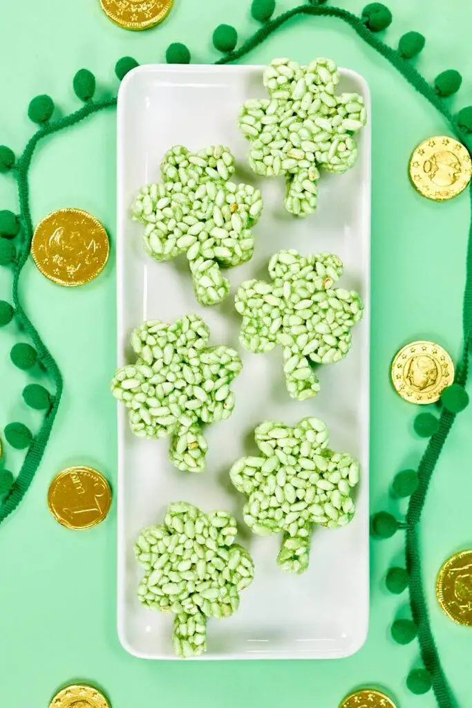 Shamrock-shaped green rice crispy treats arranged on a white tray, surrounded by gold chocolate coins and festive green decorations, perfect for St. Patrick's Day celebrations.