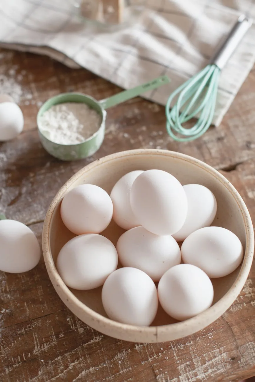 A bowl of fresh white eggs on a rustic wooden surface with baking ingredients like flour and a whisk, highlighting the need for egg substitutes in cooking and baking.