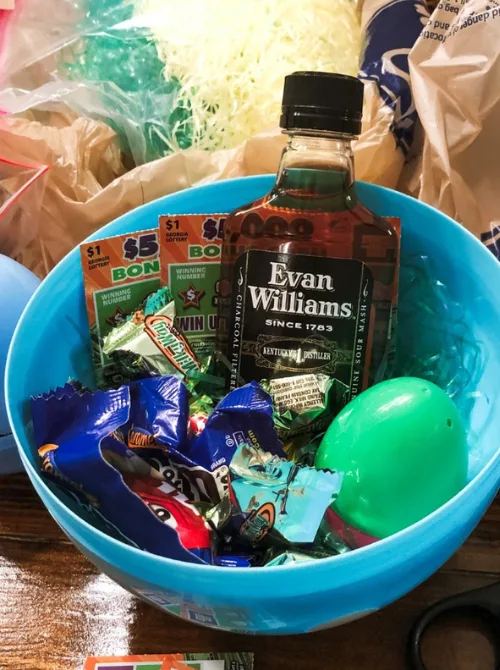 "A creative adult-themed Easter basket featuring a small bottle of Evan Williams bourbon, scratch-off lottery tickets, assorted chocolates, and a plastic Easter egg, all arranged inside a blue plastic Easter basket with festive filler. This fun and unique gift idea is perfect for those looking for DIY Easter basket ideas tailored to adults, adding a touch of excitement and indulgence to the holiday celebration.