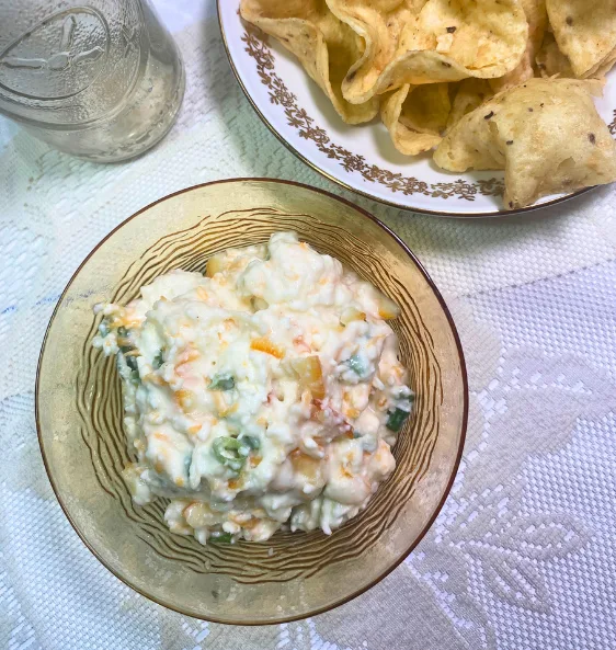 Bowl of creamy apricot jalapeño popper dip with visible green onions and shredded cheese, served with a side plate of tortilla chips on a vintage tablecloth. Perfect for game day snacks or easy party appetizers.