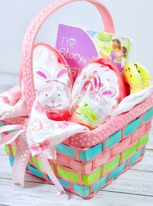 A beautifully arranged pastel Easter basket featuring a mix of thoughtful and festive gifts, including a children's book titled The Story of Easter, bunny-themed plastic eggs filled with surprises, a soft baby blanket, and a cheerful chick toy. Wrapped in a pink polka-dotted ribbon and woven with soft pastel colors, this adorable basket is a perfect inspiration for DIY Easter basket ideas for babies or toddlers, offering a sweet and meaningful way to celebrate the holiday.