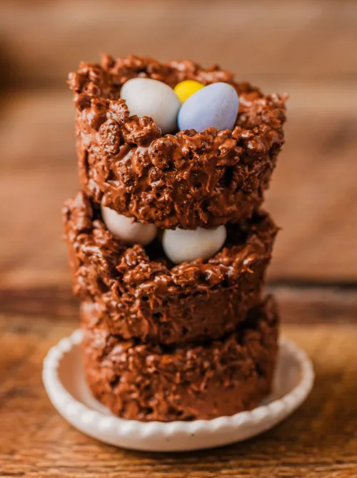 A stack of rich, chocolatey Easter nest treats made from crispy rice cereal and melted chocolate, each filled with pastel candy-coated chocolate eggs. These homemade, no-bake goodies are displayed on a rustic wooden surface, adding to their cozy and festive charm. Perfect for DIY Easter basket ideas, these sweet and crunchy nests make a delightful homemade gift or holiday treat.