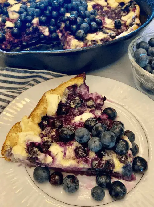 A slice of blueberry skillet pancake topped with fresh blueberries, served on a white plate with a skillet and bowl of blueberries in the background, perfect for a budget-friendly Easter brunch.