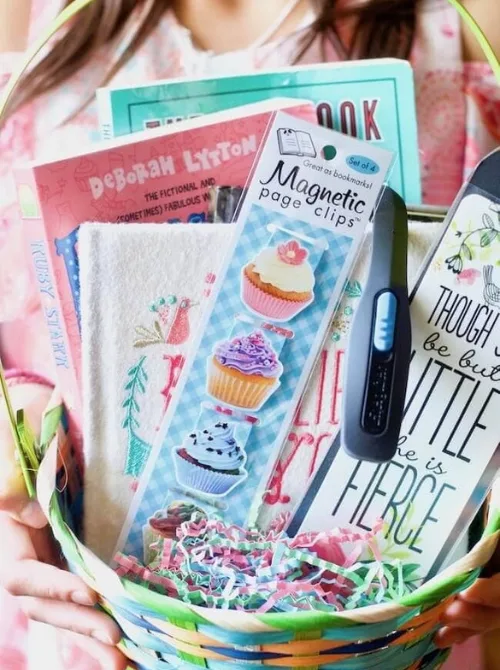 A beautifully curated book-themed Easter basket filled with thoughtful gifts, including novels, magnetic page clips with cupcake designs, a stylish bookmark, a book light, and a cozy embroidered towel. Nestled in colorful shredded paper, this unique Easter basket is perfect for book lovers, making it an inspiring addition to DIY Easter basket ideas that go beyond candy and treats.