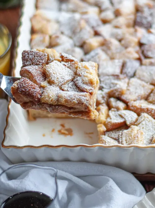 A slice of baked French toast casserole dusted with powdered sugar being lifted from a white scalloped dish—an easy and cozy option for a budget-friendly Easter brunch.