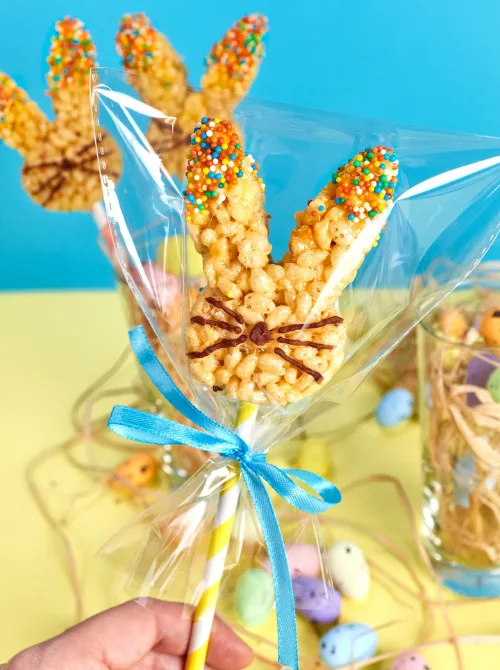 A fun and festive bunny-shaped Rice Krispies treat on a stick, decorated with colorful sprinkles on the ears and chocolate whiskers, wrapped in clear cellophane and tied with a bright blue ribbon. The treat is displayed against a cheerful pastel background with Easter eggs and shredded paper, making it a perfect addition to DIY Easter basket ideas, offering a homemade, adorable, and delicious holiday treat.