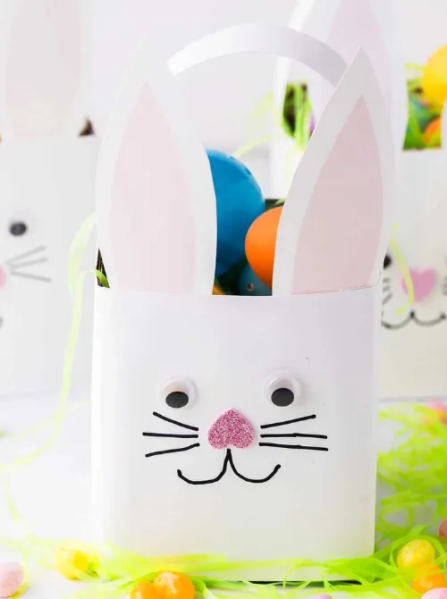 "A creative DIY bunny-themed Easter basket made from a white container, featuring adorable pink and white paper ears, googly eyes, a glittery pink nose, and a hand-drawn whiskered smile. Filled with colorful plastic eggs and surrounded by bright Easter grass, this fun and festive design is a perfect inspiration for unique Easter basket ideas that are both playful and budget-friendly.