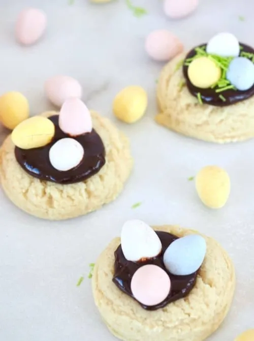 Adorable homemade Easter nest cookies featuring soft, golden-brown sugar cookies topped with a layer of rich chocolate ganache and pastel candy eggs. A sprinkle of green sugar adds a festive, grassy touch, creating a charming springtime treat. These delicious and easy-to-make cookies are a perfect addition to DIY Easter basket ideas, adding a homemade and festive flair to holiday celebrations.