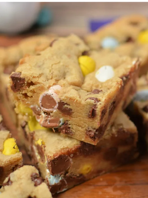 Thick and chewy Easter cookie bars loaded with chocolate chips and pastel candy-coated chocolate eggs, stacked for a delicious close-up. The golden brown bars have a soft, gooey texture with festive pops of color, making them a perfect homemade treat. These sweet and festive bars are a great addition to DIY Easter basket ideas, offering a homemade, giftable dessert that’s sure to delight.