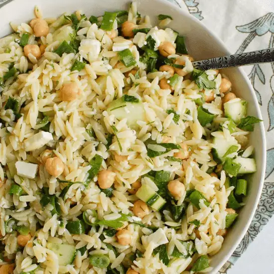 A refreshing farm to table orzo salad, packed with crisp cucumbers, chickpeas, fresh herbs, feta cheese, and a light dressing. The vibrant greens and creamy feta contrast beautifully with the tender pasta, creating a dish full of texture and flavor. A vintage spoon rests in the bowl, ready to scoop up a bite of this fresh and wholesome meal.