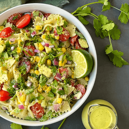 A vibrant farm to table pasta salad filled with cherry tomatoes, sweet corn, avocado, red onion, and fresh cilantro, tossed with a creamy dressing. A bright lime wedge sits on top, ready to add a zesty burst of flavor. The fresh, colorful ingredients highlight the beauty of farm to table eating—simple, wholesome, and full of delicious, seasonal goodness.