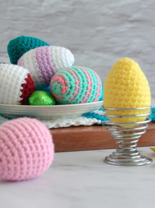 A collection of beautifully handcrafted crochet Easter eggs in pastel and vibrant colors, displayed in a dish and a metal egg holder. Each egg features unique patterns and textures, adding a cozy, handmade charm to Easter decor. These soft, reusable eggs make a wonderful addition to DIY Easter basket ideas, offering a creative, eco-friendly, and long-lasting alternative to traditional plastic eggs.S