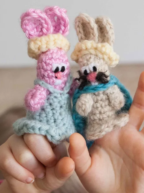 A pair of adorable handmade crochet bunny finger puppets, dressed in tiny blue and beige outfits with charming details like little hats and embroidered faces. The pink and beige bunnies are held on a child's fingers, bringing them to life for playful storytelling. These cute and creative toys are a wonderful addition to DIY Easter basket ideas, adding a handmade, heartfelt touch to holiday gifts.