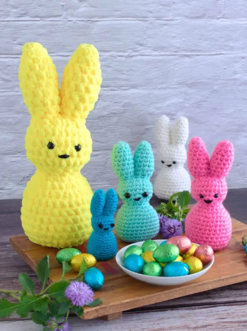 A delightful collection of handmade crochet bunnies in bright pastel colors, including yellow, pink, blue, and white, arranged alongside a dish of foil-wrapped chocolate eggs and fresh greenery. With their soft, textured design and adorable facial details, these cuddly bunnies make a charming addition to DIY Easter basket ideas, offering a handmade and heartfelt touch to holiday gifting.