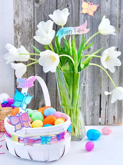 A beautifully decorated white Easter basket filled with colorful plastic eggs, adorned with pastel butterfly decorations, sitting beside a vase of fresh white tulips on a rustic wooden backdrop. A 'Happy Easter' sign adds a festive touch. A perfect inspiration for unique Easter baskets with a delicate spring theme.