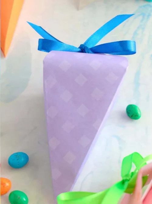 A beautifully crafted DIY carrot-shaped Easter treat box made from pastel purple patterned paper, tied with a bright blue ribbon. Surrounded by colorful candy eggs and festive decorations, this handmade gift box is a creative and fun addition to DIY Easter basket ideas, perfect for filling with small treats or surprises for a personalized holiday touch.