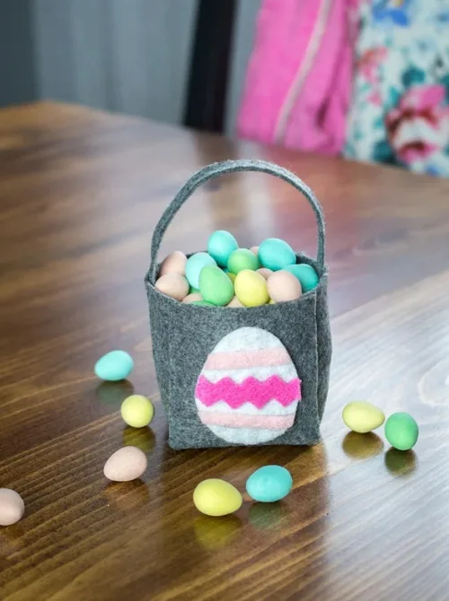 A small, handcrafted gray felt Easter basket with a cute pink and white egg design on the front, filled with pastel-colored candy eggs. The basket sits on a wooden table with more candy eggs scattered around, creating a festive and playful scene. This DIY-inspired design is perfect for unique Easter basket ideas, offering a charming and minimalist approach to holiday gifting.