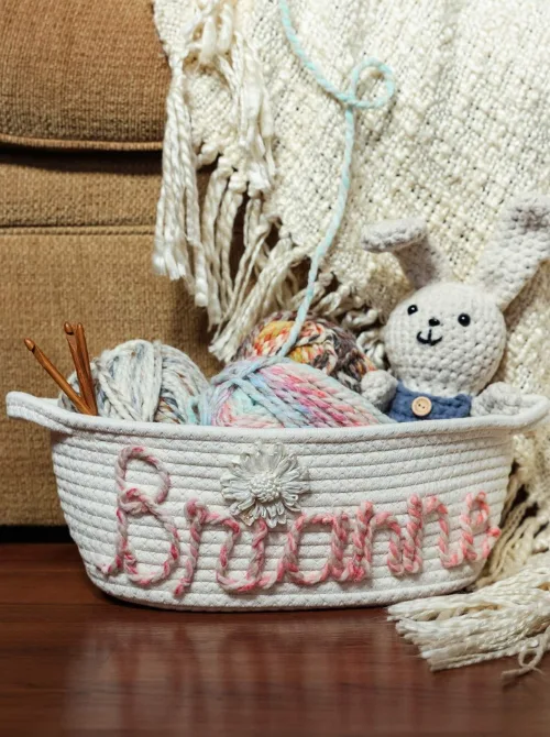 A personalized rope basket with the name 'Brianna' stitched in soft pink yarn, filled with pastel-colored yarn, crochet hooks, and a handmade crocheted bunny wearing a blue outfit. The cozy setup is placed near a fringed cream-colored blanket, creating a warm and rustic feel. This thoughtful and handcrafted design is a perfect inspiration for unique Easter basket ideas, especially for craft lovers and those who appreciate DIY gifts with a personal touch.