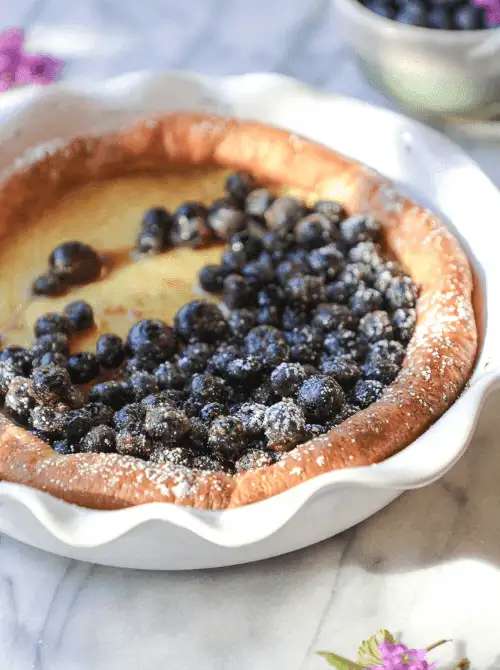 Golden baked Dutch baby pancake topped with powdered sugar and fresh blueberries, served in a white scalloped dish—perfect for a budget-friendly Easter brunch.