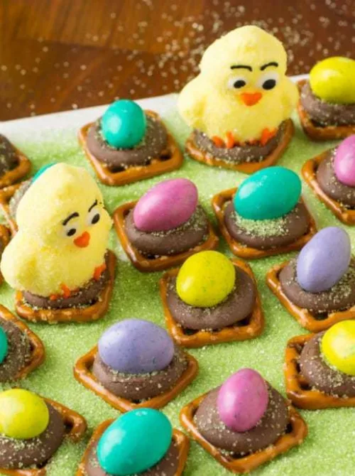 "A festive Easter treat display featuring bite-sized chocolate and pretzel nests topped with pastel candy eggs, along with adorable marshmallow chicks perched on some of the nests. The treats are sprinkled with green sugar for a playful, grass-like effect, creating a fun and delicious Easter snack. These easy-to-make sweets are a perfect addition to DIY Easter basket ideas, adding a homemade and whimsical touch to holiday celebrations.