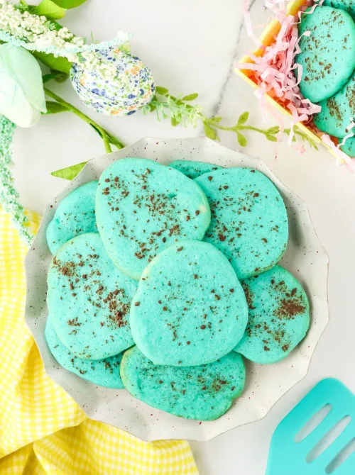 A plate of vibrant blue egg-shaped Easter cookies with a speckled chocolate dusting, resembling robin’s eggs. The cookies are beautifully displayed alongside festive spring decorations, a yellow gingham cloth, and a basket filled with more cookies nestled in pastel Easter grass. These creative and eye-catching treats make a delightful addition to DIY Easter basket ideas, offering a homemade and festive touch to holiday celebrations.