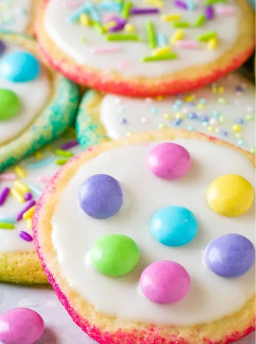 Colorful Easter sugar cookies decorated with white icing, pastel candy-coated chocolates, and festive sprinkles. The cookies have a fun, vibrant design with a soft gradient of pink and yellow around the edges, adding to their festive charm. These sweet treats make a delightful addition to DIY Easter basket ideas, offering a homemade and delicious way to celebrate the holiday.