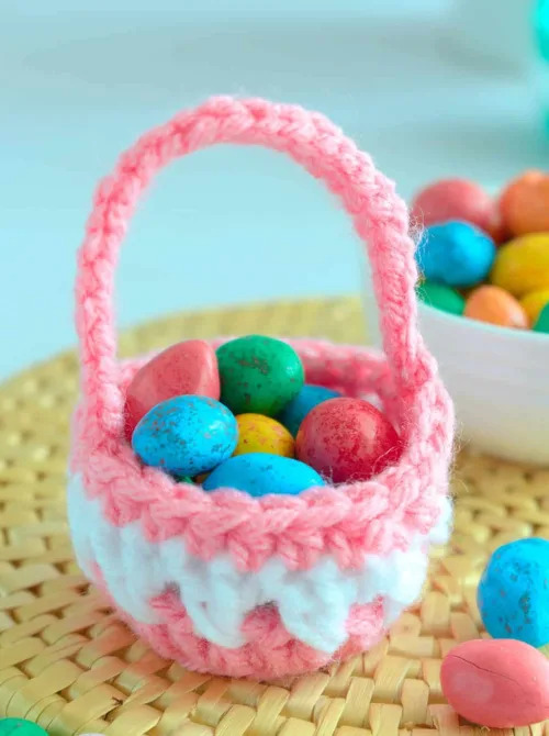 A charming handmade crochet Easter basket in soft pink and white, filled with colorful speckled candy eggs. The tiny basket, crafted with intricate stitches and a sturdy handle, sits on a woven placemat, adding a cozy and festive touch. This adorable DIY creation is perfect for DIY Easter basket ideas, offering a unique and reusable way to gift sweet treats for the holiday.