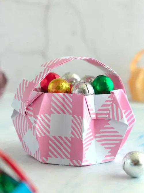 A charming DIY Easter basket made from pink and white plaid patterned paper, carefully folded into a unique geometric design with a matching handle. Filled with colorful foil-wrapped chocolate eggs, this handmade creation is a perfect inspiration for DIY Easter basket ideas, offering a fun and budget-friendly way to package sweet treats for the holiday.