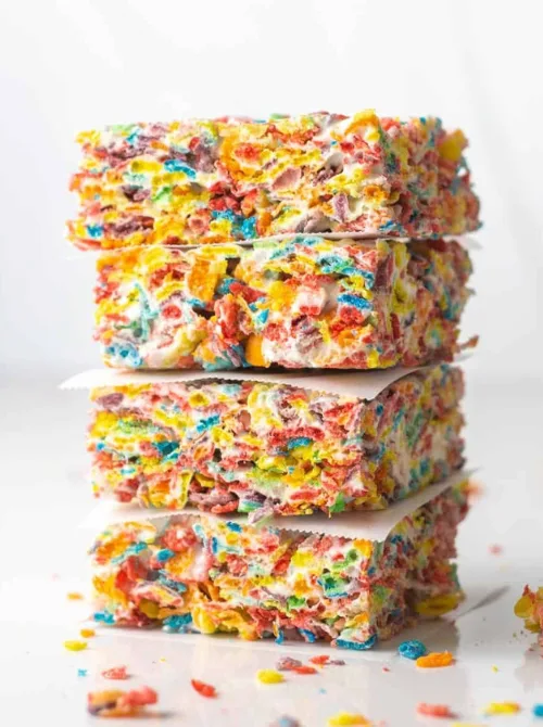 A vibrant stack of Fruity Pebbles marshmallow treats, bursting with bright colors and gooey, melted marshmallow goodness. These no-bake cereal bars are cut into perfect squares and layered with parchment paper for a neat presentation. A fun and festive addition to DIY Easter basket ideas, these sweet and colorful treats make a delightful homemade gift or holiday snack.