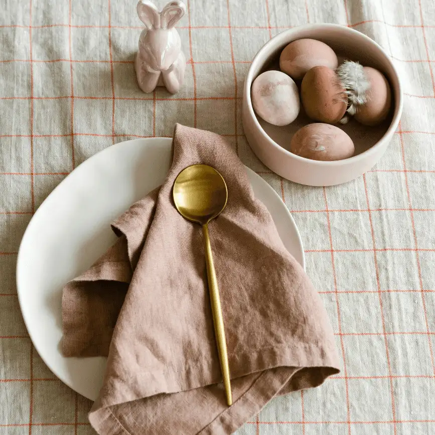How to Plan a Surprisingly Budget-Friendly Easter Brunch Minimalist Easter brunch table setting with a white plate, blush pink cloth napkin, gold spoon, bowl of brown eggs, and a small pink bunny figurine on a neutral plaid tablecloth.