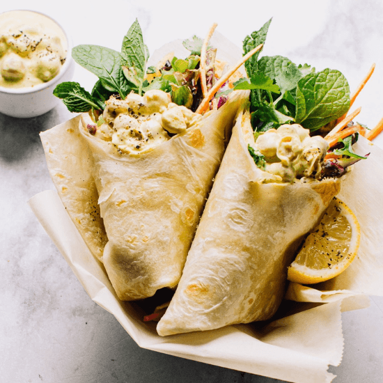 A pair of hearty farm to table wraps, filled with a creamy chickpea salad, fresh greens, shredded carrots, and fragrant herbs, all tucked inside soft, golden flatbreads. A lemon wedge sits beside them, ready to add a burst of brightness. The natural, locally sourced ingredients make these wraps both nourishing and flavorful, perfect for a fresh and wholesome meal.