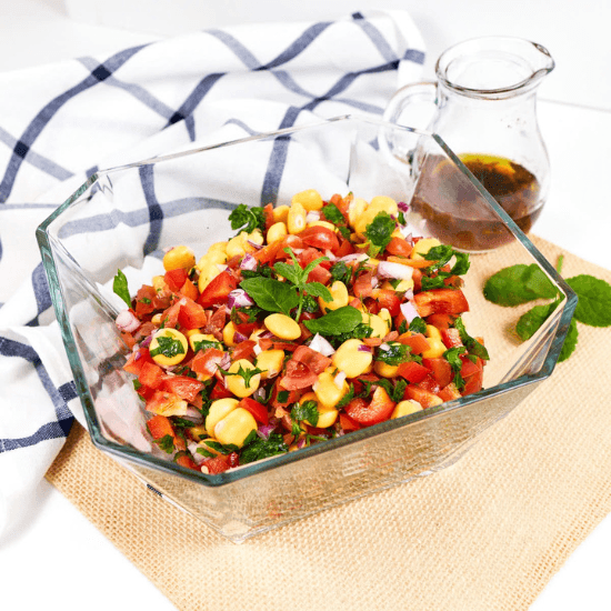 A fresh and colorful farm to table chickpea salad, featuring juicy tomatoes, crisp red bell peppers, red onion, and fragrant herbs, all tossed in a light vinaigrette. Served in a clear glass bowl, the vibrant ingredients shine, highlighting the natural beauty of locally sourced produce. A small pitcher of dressing sits nearby, ready to enhance the bright, refreshing flavors.