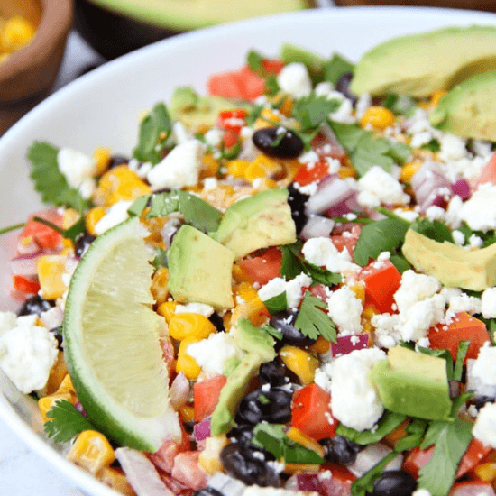 A vibrant farm-to-table salad bursting with fresh ingredients, including black beans, roasted corn, diced tomatoes, red onion, avocado slices, crumbled cheese, and chopped cilantro. A fresh lime wedge rests on top, ready to add a zesty kick. The dish showcases the rich flavors and colors of locally sourced produce, emphasizing freshness and wholesome goodness.