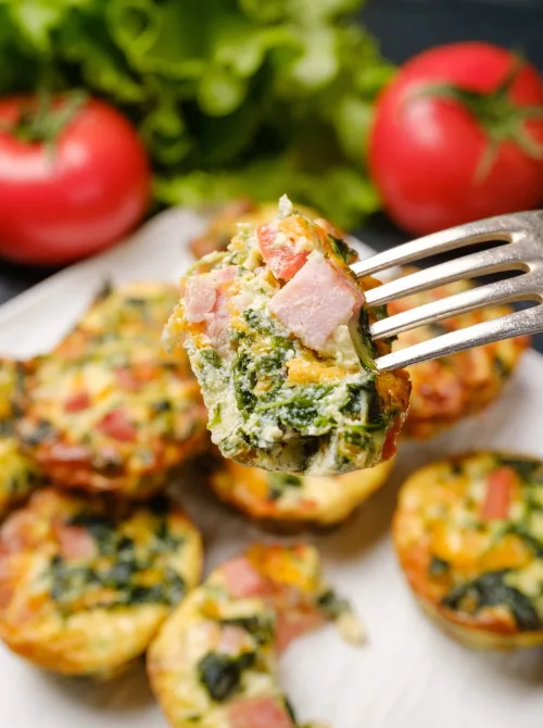 Close-up of a fork holding a bite-sized ham, spinach, and cheese egg muffin with fresh tomatoes and greens in the background—perfect for a budget-friendly Easter brunch.