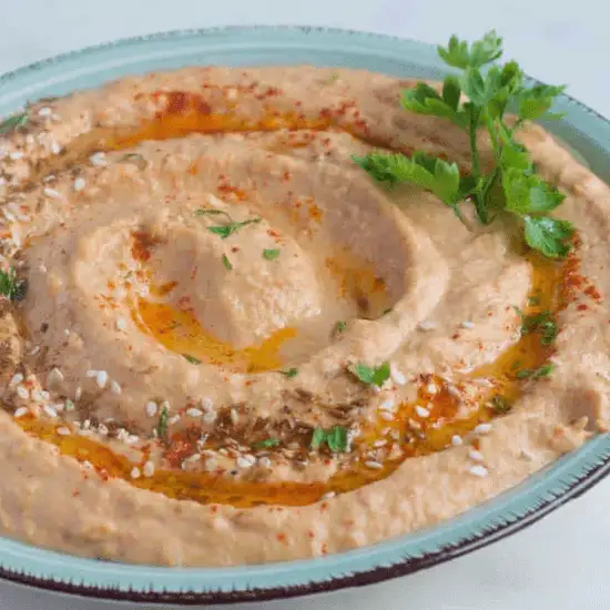 A creamy bowl of farm to table hummus, drizzled with golden olive oil and sprinkled with paprika, sesame seeds, and fresh herbs. The smooth texture and rich color highlight the freshness of locally sourced ingredients, creating a wholesome and flavorful dish. A sprig of parsley adds a final touch, making this hummus both visually inviting and deliciously nourishing.