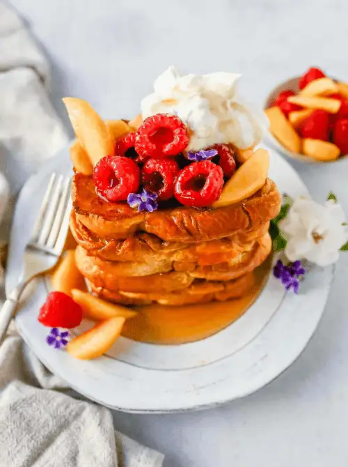 Stack of fluffy French toast topped with fresh raspberries, peach slices, syrup, and whipped cream, beautifully garnished for a budget-friendly Easter brunch.