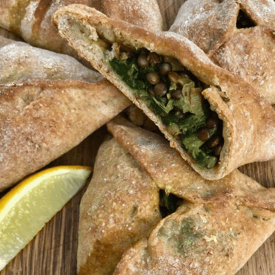 A batch of golden-brown farm to table parcels filled with tender lentils and fresh greens, baked in a rustic whole wheat dough. One pastry is cut open, revealing the hearty, nutrient-packed filling. A wedge of lemon sits nearby, ready to add a bright, zesty touch. The wholesome ingredients and handmade feel highlight the essence of farm-to-table cooking—fresh, simple, and full of flavor.