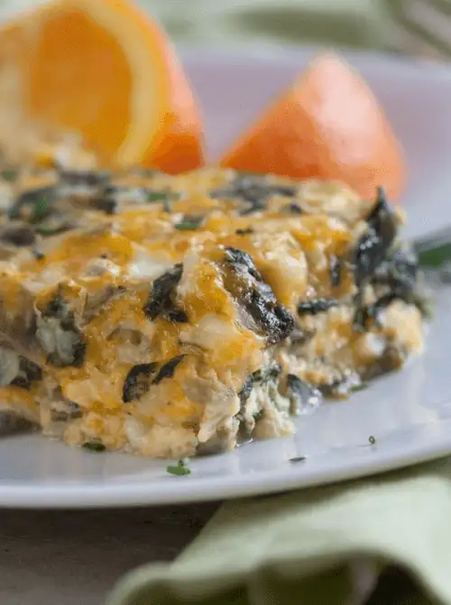 Close-up of a cheesy spinach and mushroom breakfast casserole served on a white plate with orange wedges—hearty and perfect for a budget-friendly Easter brunch.