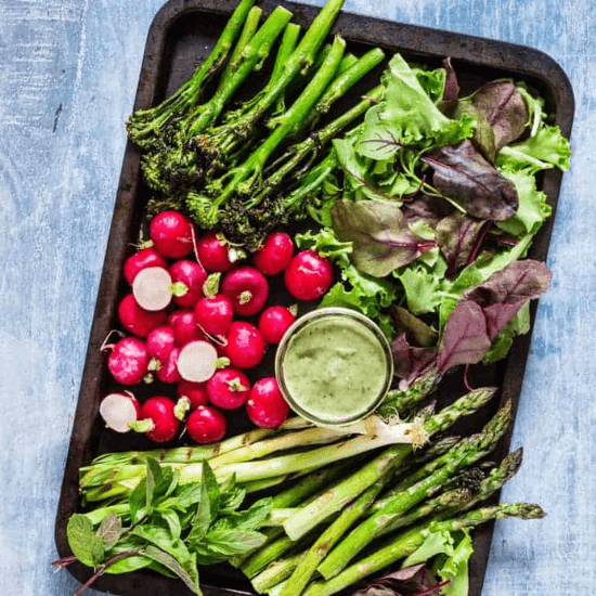 A beautifully arranged tray of farm to table fresh vegetables, including charred broccolini, crisp radishes, tender asparagus, leafy greens, and fragrant herbs. A small bowl of creamy green dressing sits in the center, ready for dipping. The vibrant colors and fresh-picked quality highlight the essence of farm to table eating—seasonal, nutritious, and bursting with flavor.