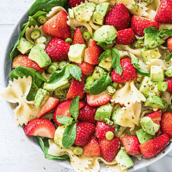 A refreshing farm to table pasta salad featuring sweet strawberries, creamy avocado, fresh mint, and tender bowtie pasta, all tossed with a light poppy seed dressing. The vibrant colors and fresh, seasonal ingredients create a perfect balance of flavors and textures, making this dish a delightful blend of sweet and savory goodness.