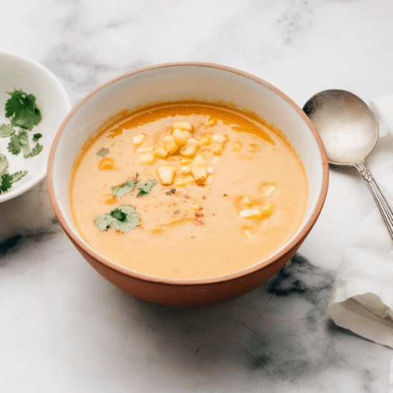 A comforting bowl of farm to table creamy sweet potato and corn soup, garnished with sweet corn kernels, fresh cilantro, and a hint of red pepper flakes. The rich golden color and velvety texture highlight the freshness of locally sourced ingredients, making this dish a warm and nourishing delight perfect for any season.