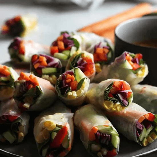 A plate of fresh farm-to-table spring rolls, packed with vibrant vegetables like red bell peppers, carrots, cucumbers, purple cabbage, and leafy greens, wrapped in delicate rice paper. A small bowl of dipping sauce sits beside them, ready for dipping. The natural light highlights the crisp, just-picked produce, emphasizing the freshness and quality of locally sourced ingredients.