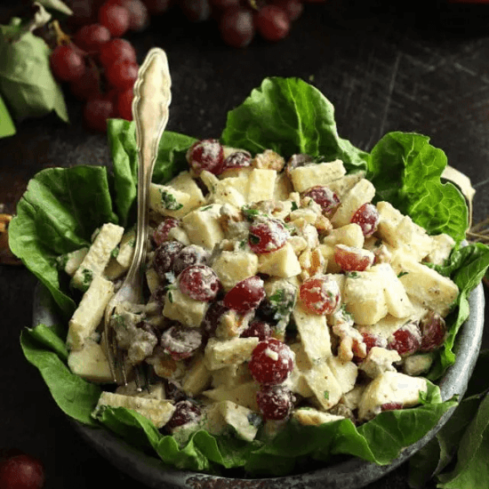 A fresh and vibrant farm to table Waldorf salad, featuring crisp apples, juicy red grapes, crunchy walnuts, and a creamy dressing, all served over a bed of leafy green lettuce. A vintage fork rests in the bowl, ready for a bite. The rich colors and wholesome ingredients highlight the beauty of farm to table eating—simple, fresh, and full of natural goodness.S