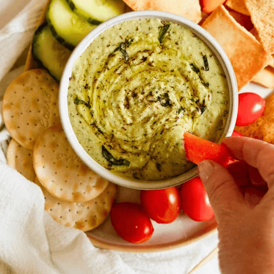 A creamy bowl of farm to table dip, swirled with fresh herbs and drizzled with olive oil, served alongside crisp cucumbers, cherry tomatoes, crackers, and warm pita. A hand dips a vibrant red bell pepper into the smooth, flavorful spread, highlighting the freshness of locally sourced ingredients. This wholesome, colorful platter embodies the simplicity and richness of farm to table eating.